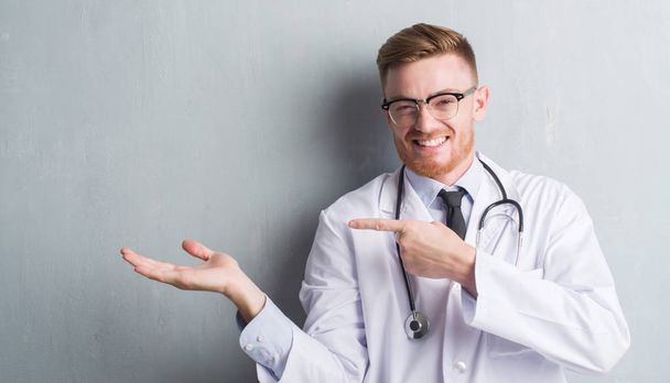 Young redhead doctor man over grey grunge wall very happy pointing with hand and finger - Фото, изображение