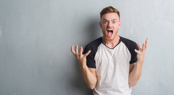 Young redhead man over grey grunge wall crazy and mad shouting and yelling with aggressive expression and arms raised. Frustration concept. - Photo, Image