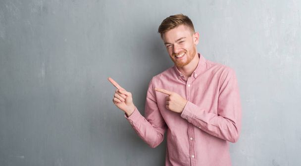 Young redhead man over grey grunge wall wearing pink shirt smiling and looking at the camera pointing with two hands and fingers to the side. - Photo, Image