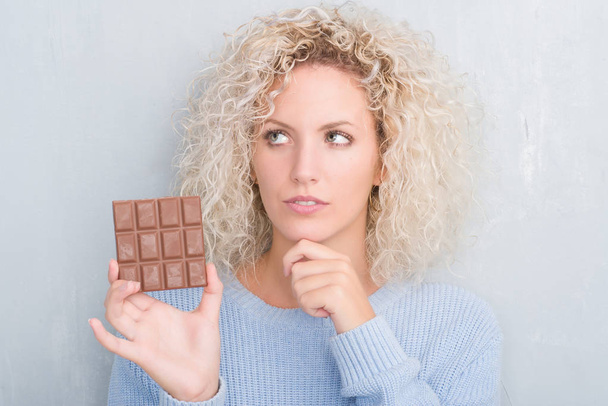 Mladá blondýnka nad grunge šedá zeď jíst čokoláda vážná tvář přemýšlet o otázku, velmi zmatené myšlenky - Fotografie, Obrázek