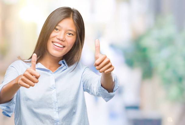 Young asian business woman over isolated background approving doing positive gesture with hand, thumbs up smiling and happy for success. Looking at the camera, winner gesture. - Foto, Imagem