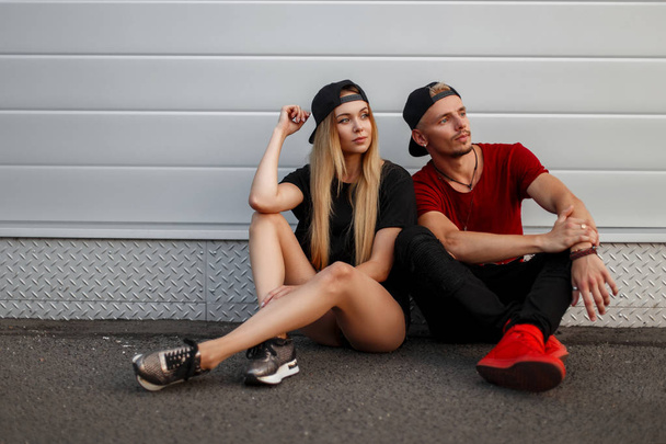 Όμορφο νεαρό ζευγάρι μόδας σε ρούχα κομψά καλοκαίρι κάθεται κοντά σε ένα τοίχο στο δρόμο - Φωτογραφία, εικόνα