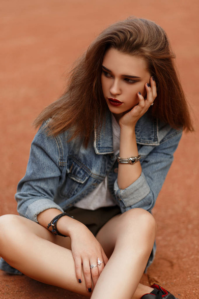 Beautiful stylish woman model in a denim jacket with shorts sitting on the sand in the park - Photo, Image