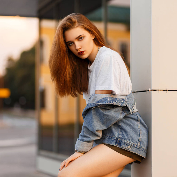 Stylish beautiful sexy model woman in trendy clothes with a jeans jacket posing outdoors near a building - Fotó, kép