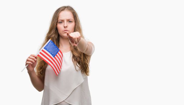 Giovane donna bionda che tiene la bandiera degli Stati Uniti punta con il dito alla fotocamera e a te, segno della mano, gesto positivo e fiducioso dal davanti
 - Foto, immagini