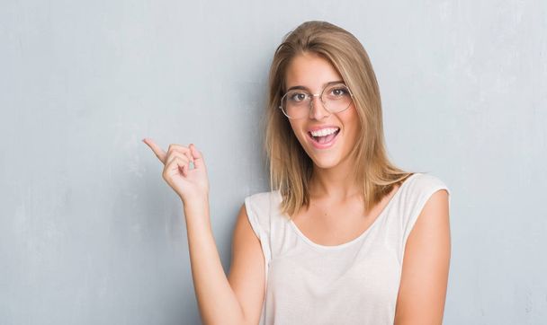 Hermosa mujer joven de pie sobre la pared gris grunge con gafas muy felices señalando con la mano y el dedo a un lado
 - Foto, imagen