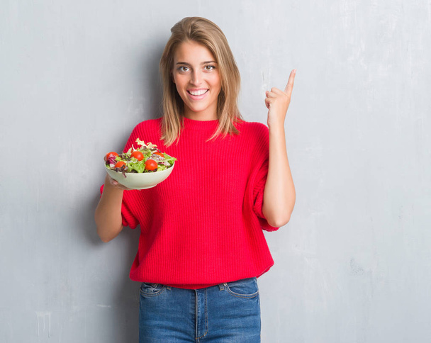 Beautiful young woman over grunge grey wall eating tomato salad surprised with an idea or question pointing finger with happy face, number one - Photo, Image