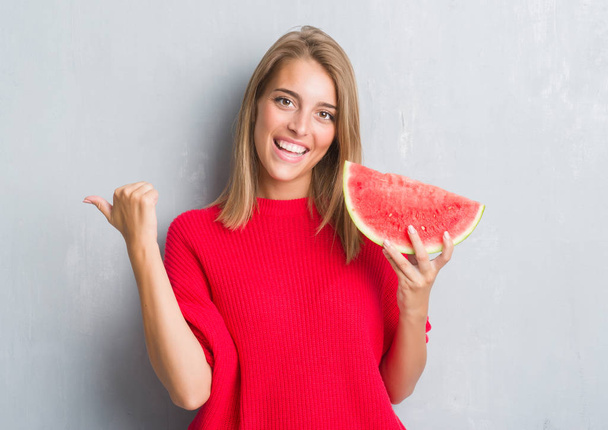Mooie jonge vrouw over grunge grijs muur eten water meloen aan te wijzen en te tonen met de duim aan de kant met een blij gezicht lachend - Foto, afbeelding