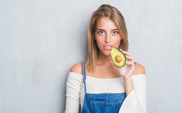 Красива молода жінка над гранжевою сірою стіною їсть авокадо з впевненим виразом на розумне обличчя думаючи серйозно
 - Фото, зображення