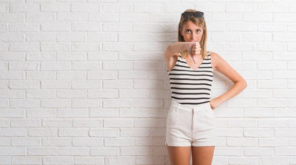 Young beautiful blonde woman over white brick wall looking unhappy and angry showing rejection and negative with thumbs down gesture. Bad expression. - Photo, image