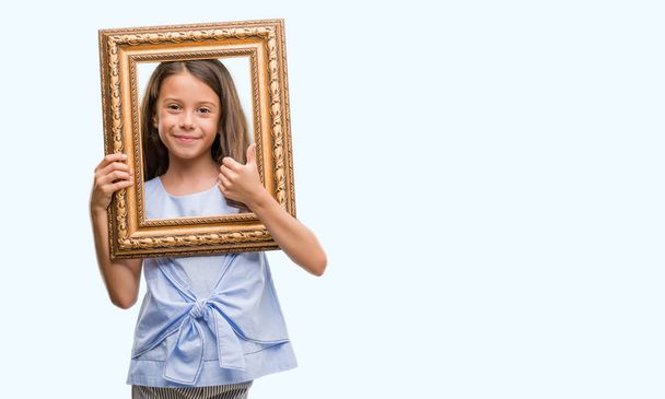 Brunette hispanic girl holding vintage art frame happy with big smile doing ok sign, thumb up with fingers, excellent sign - Фото, изображение