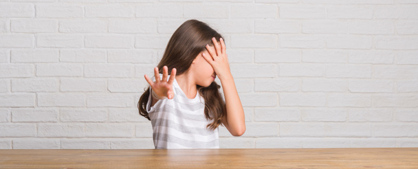 Молода іспаномовна дитина сидить на столі вдома, прикриваючи очі руками і роблячи стоп жест з сумним і страховим виразом. Збентежена і негативна концепція
. - Фото, зображення