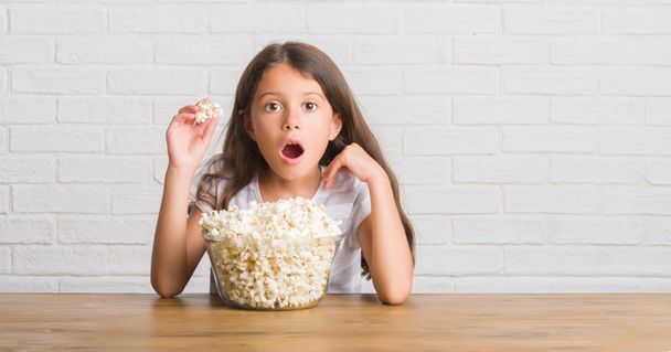 Young hispanic kid sitting on the table eating pop corn scared in shock with a surprise face, afraid and excited with fear expression - Foto, imagen