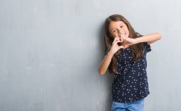 Młode dziecko hiszpanin streszczenie szary mur uśmiechający się w miłość serce symbol i kształt rękami. Romantyczna koncepcja. - Zdjęcie, obraz