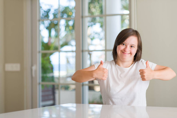 Down syndrome woman at home success sign doing positive gesture with hand, thumbs up smiling and happy. Looking at the camera with cheerful expression, winner gesture. - Photo, Image