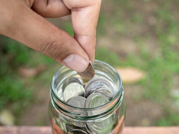 Close-up hand holding coin and put in glass jar on wooden table and on green tree background. Saving money concept. - Photo, Image