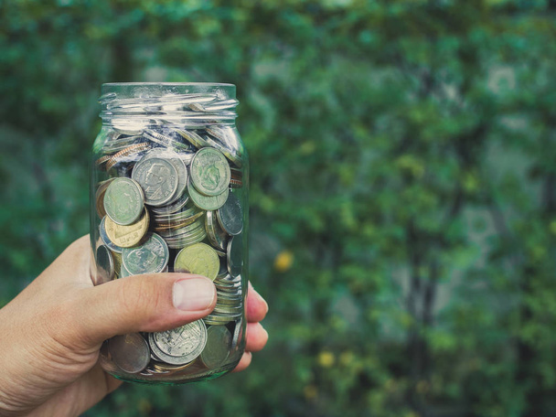 Рука держит монеты в стеклянной банке на зеленом фоне дерева с копировальным пространством. Экономия денег
. - Фото, изображение