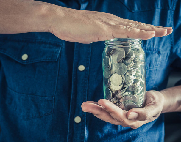 Man wearing blue shirt with hands holding coins in glass jar. Saving money and donation concept. - Photo, Image