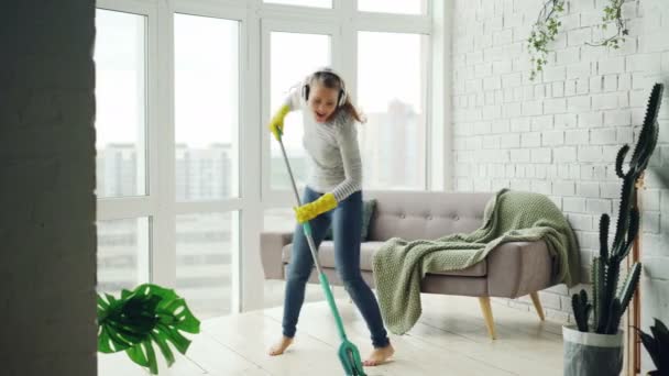 Happy housewife is dancing and singing during housecleaning, she is listening to music in headphones and mopping floor having fun. Housework and technology concept. - Video