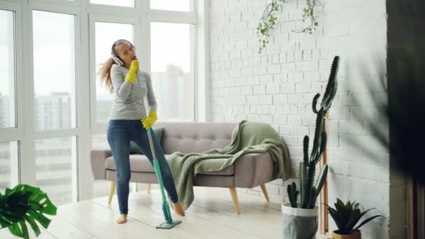 Joyful girl is mopping floor in light apartment and having fun listening to music through headphones, dancing and singing. Beautiful furniture and plants is visible. - Кадры, видео