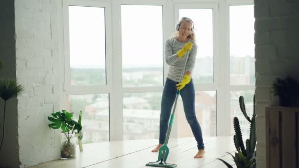 Beautiful young woman is cleaning light modern studio with panoramic windows, girl is washing floor and having fun listening to music, singing and dancing. - Séquence, vidéo