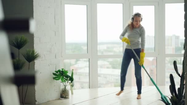 Joyful young lady is doing housework mopping floor and enjoying music in headphones, dancing and singing. Beautiful apartment with green plants is visible. - Video