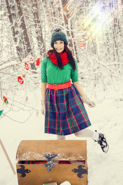 brunette girl in a green hat and in a costume of flowers traditional to the elves Santas helpers in the winter forest under the snow with a chest of gifts and giant candy - Photo, Image