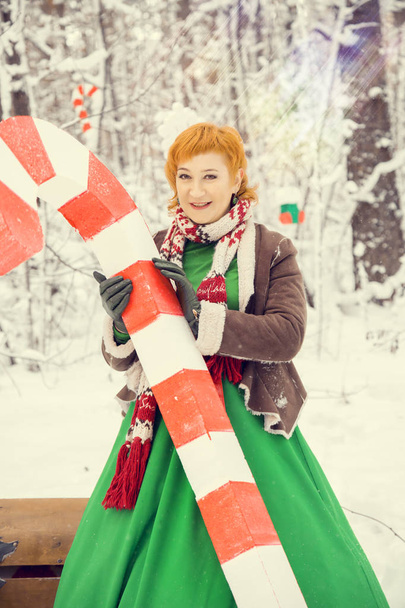 femme rousse ardente dans une robe de bal verte avec une ceinture en cuir rouge dans le costume d’assistant nain Santa Claus dans la forêt en hiver avec bonbons énormes, une poitrine de cadeaux, le concept de la nouvelle année - Photo, image