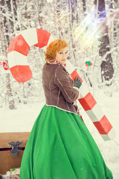 fiery red-haired woman in a ball green dress with a red leather belt in the costume of dwarf assistant Santa Claus in the winter forest with huge candy, a chest of gifts, the concept of the new year - Valokuva, kuva