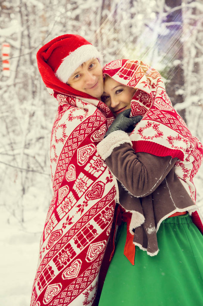 young couple, man and woman, husband and wife are walking in costumes of flowers typical of the elves of Santas helpers in a winter forest under the snow with a chest full of gifts and a giant candy - Photo, Image