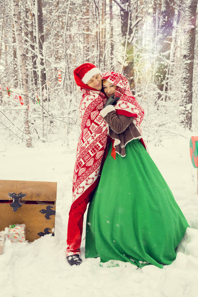 young couple, man and woman, husband and wife are walking in costumes of flowers typical of the elves of Santas helpers in a winter forest under the snow with a chest full of gifts and a giant candy - Photo, Image