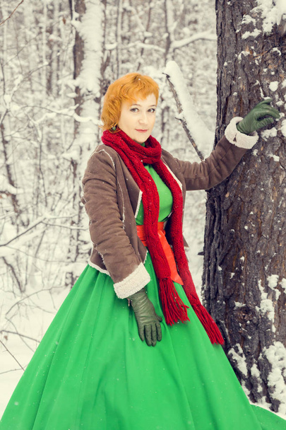 φλογερή κοκκινομάλλα γυναίκα σε ένα φόρεμα μπάλα πράσινη με κόκκινη δερμάτινη ζώνη με το κοστούμι του νάνος βοηθό Άγιου Βασίλη μέσα στο δάσος του χειμώνα με τεράστια καραμέλα, ένα στήθος των δώρων, η έννοια του νέου έτους - Φωτογραφία, εικόνα