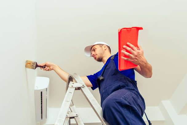 Όμορφος εργάτης στην Σκάλα ζωγραφική τοίχου σε λευκό χρώμα με πινέλο - Φωτογραφία, εικόνα