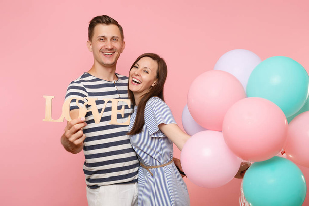 Portrait couple hold wooden word letters love. Woman and man in blue clothes celebrating birthday holiday party on pastel pink background with colorful air balloons. People sincere emotions concept - Valokuva, kuva