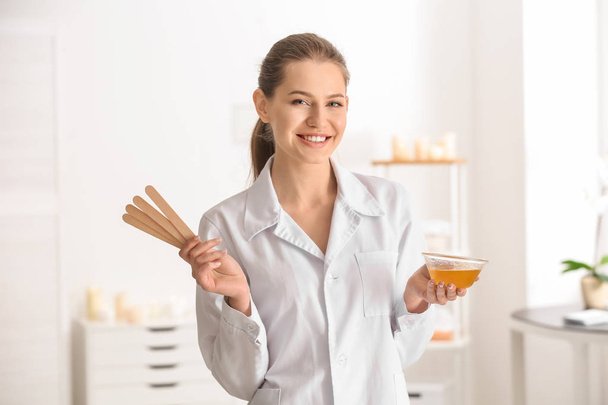 Vrouwelijke schoonheidsspecialist holding sticks voor ontharing en kom met wax op onscherpe achtergrond - Foto, afbeelding