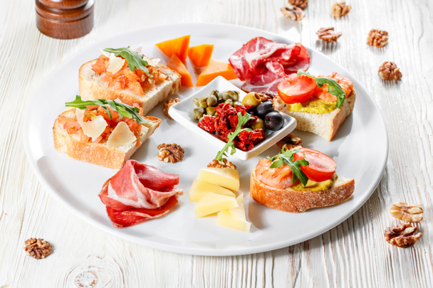 Plate with Italian appetizers. Bruschetta with a cherry tomatoes and shrimps. Parmesan cheese, prosciutto, green capers, olives, sun-dried tomatoes and walnuts. - Photo, Image