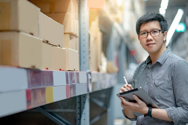 Nuori aasialainen mies työntekijä tekee tilannearvion tuotteen pahvilaatikossa hyllyillä varastossa digitaalisen tabletin ja kynän avulla. Varastonmäärityksen käsite
 - Valokuva, kuva