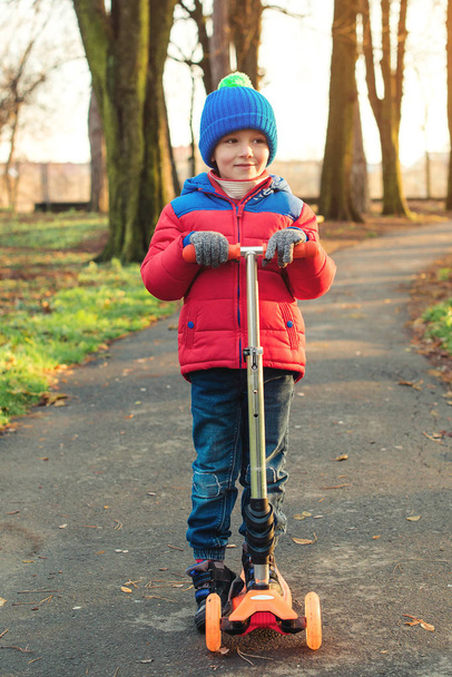 Забавный маленький мальчик катается на скутере холодной осенью в парке. Ребенок играет на улице со скутерами. Активный отдых и открытый спорт для детей. Счастливого дошкольника в красочной модной одежде. Осенняя мода
 - Фото, изображение