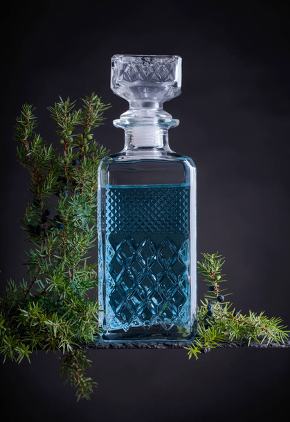 Blauer Gin in Kristallkaraffe und Wacholderzweig mit Beeren auf schwarzem Hintergrund. - Foto, Bild