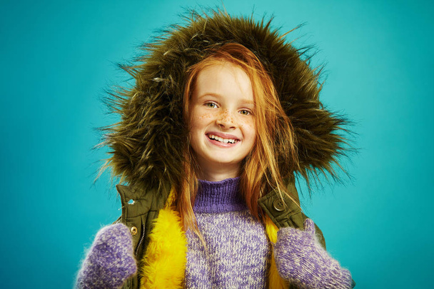 joyful girl of ten years in winter clothes over blue background. Red haired child with beautiful freckles smiles, dressed in warm jacket, enjoys winter. - Foto, Bild
