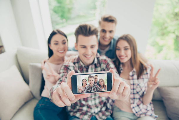 Smartphone odak bulanık bir arka plan ile dört genç insanların modern telefonunuzun ekranında tüm dikkatleri çekmek denim giysiler içinde bir kanepede oturan selfie - Fotoğraf, Görsel