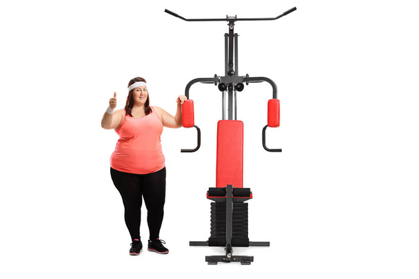 Full length portrait of an overweight woman with an exercising machine showing thumbs up isolated on white background - Photo, image