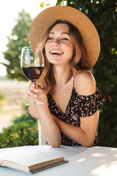Image of cute pretty young woman sitting in cafe outdors in park with book holding glass drinking wine. - Фото, изображение