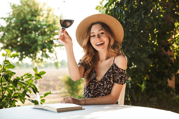 Photo of happy young woman wearing straw hat drinking wine while sitting at table and reading book outdoor in cafe - Foto, afbeelding
