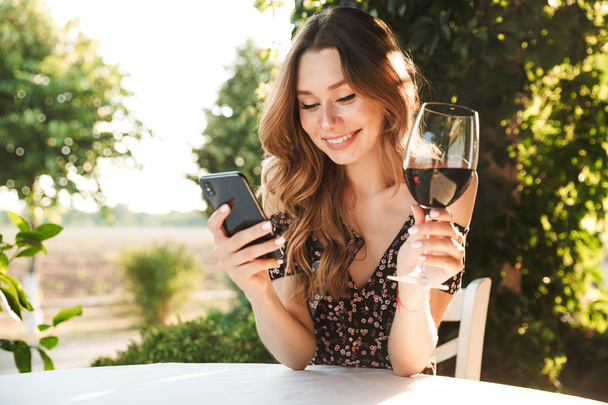 Image of young lady sitting in cafe outdors in park holding glass drinking wine chatting by mobile phone. - Фото, изображение