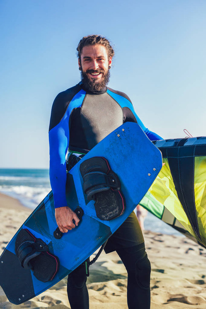 Yakışıklı beyaz adam profesyonel sörfçü onun uçurtma ve tahta ile kumlu plajda durup. - Fotoğraf, Görsel