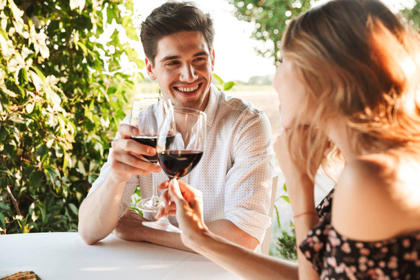 Picture of young loving couple sitting in cafe by dating outdors in park holding glasses of wine drinking. - Photo, image