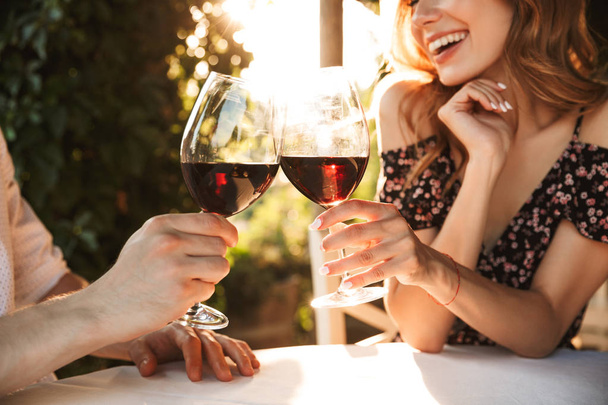 Cropped image of young loving couple sitting in cafe by dating outdors in park holding glasses of wine drinking. - Zdjęcie, obraz