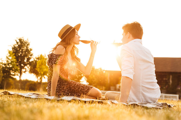 Image of young loving couple sitting by dating outdors on picnic holding glasses of wine drinking. - Foto, imagen