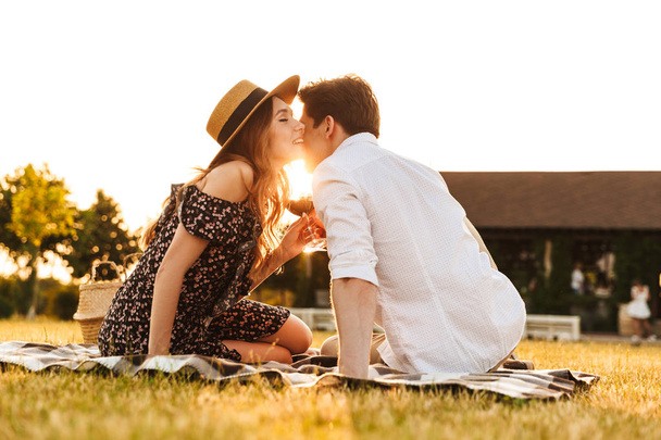 Image of young cute loving couple sitting by dating outdors on picnic holding glasses of wine kissing. - Photo, image